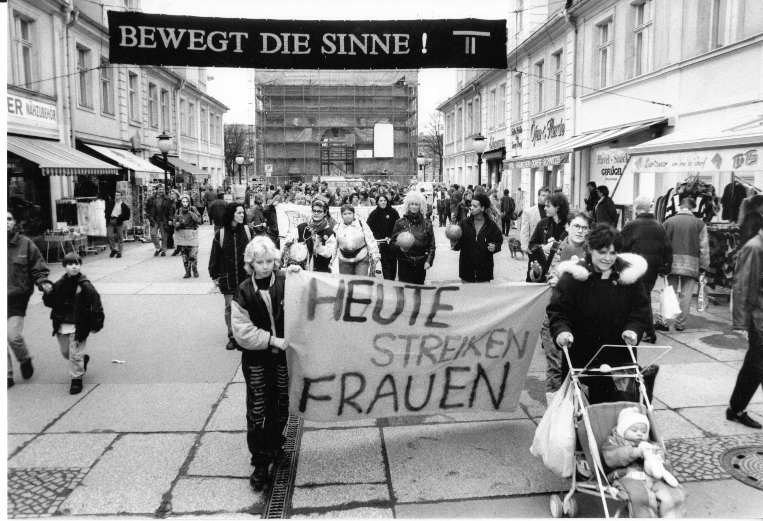 Schwarz-weiß-Fotografie einer Demonstration des Unabhängigen Frauenverbunds in der Brandenburger Straße, Berlin 1994: Frauen tragen ein Transparent mit der Aufschrift ‚Heute streiken Frauen‘, Passanten und Kinder im Vordergrund.