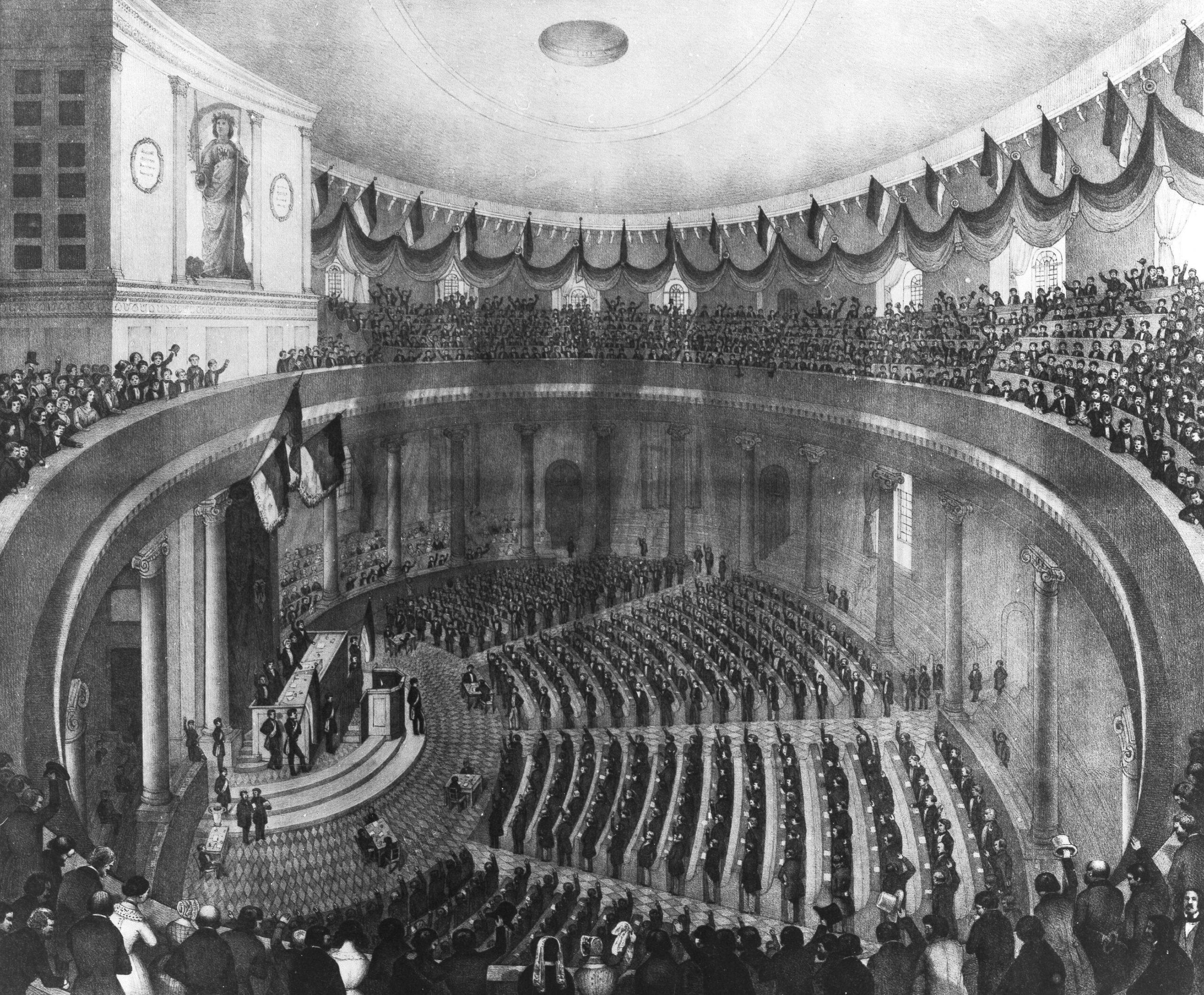 Historische Schwarz-weiß-Abbildung des Innenraums der Frankfurter Paulskirche während der Nationalversammlung 1848, mit dicht besetzten Rängen und Rednerpodium.