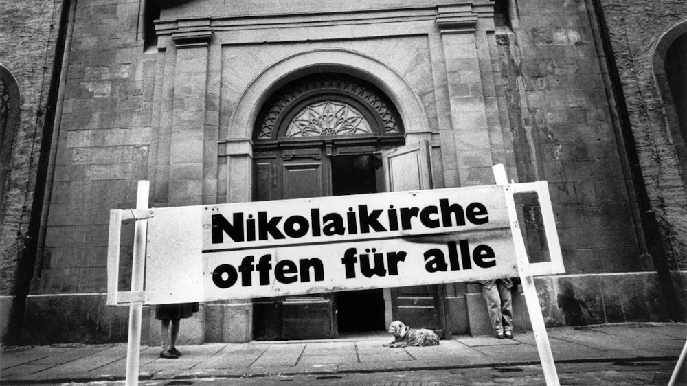 Schwarz-weiß-Fotografie des Eingangs der Nikolaikirche mit Schild "Nikolaikirche offen für alle", davor ein kleiner Hund auf der Treppe.